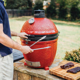 Hombre cocinando en un Kamado Joe rojo en el jardín, con carne en la tabla al lado