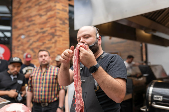 Hombre sostiene costilla de cerdo cruda durante curso de asados y parrilla, rodeado de personas.