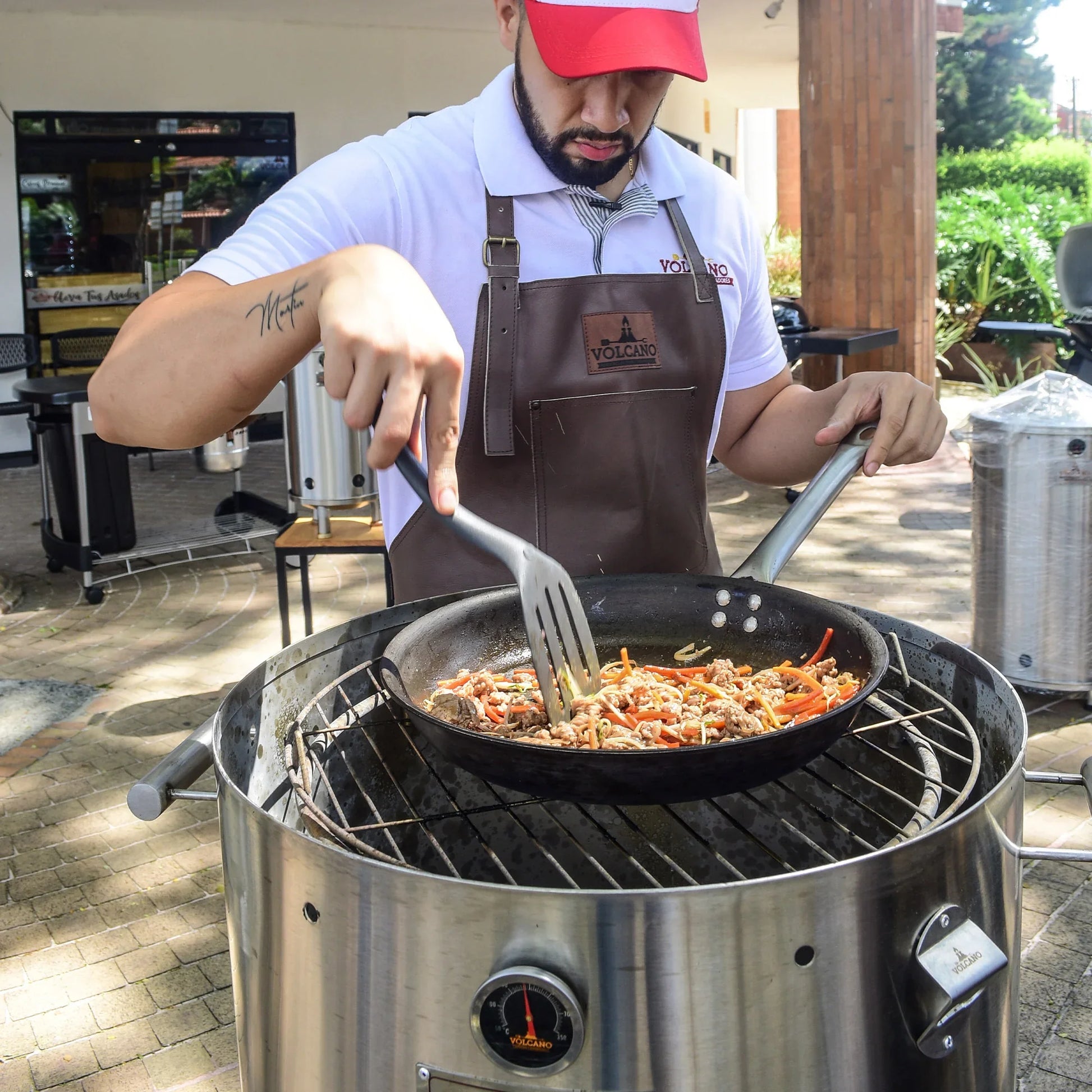 Hombre cocina carne y verduras en sartén sobre barril asador de acero inoxidable al aire libre