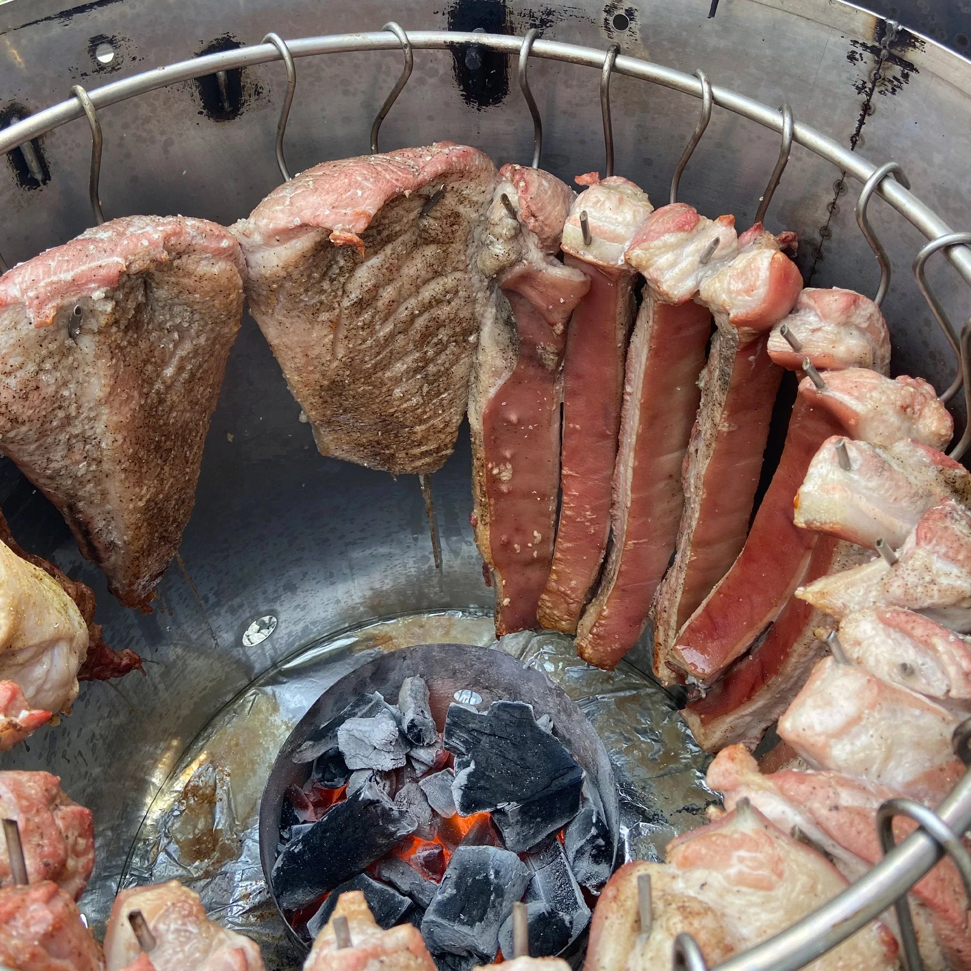 Carne colgada en barril asador jumbo de acero inoxidable con carbón encendido, ideal para asados.
